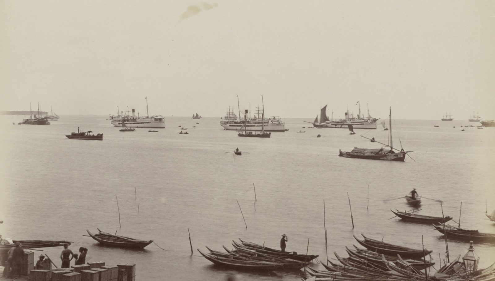 Steamships and sailing ships at anchor in Singapore harbour, with small wooden boats moored along the shore in front.