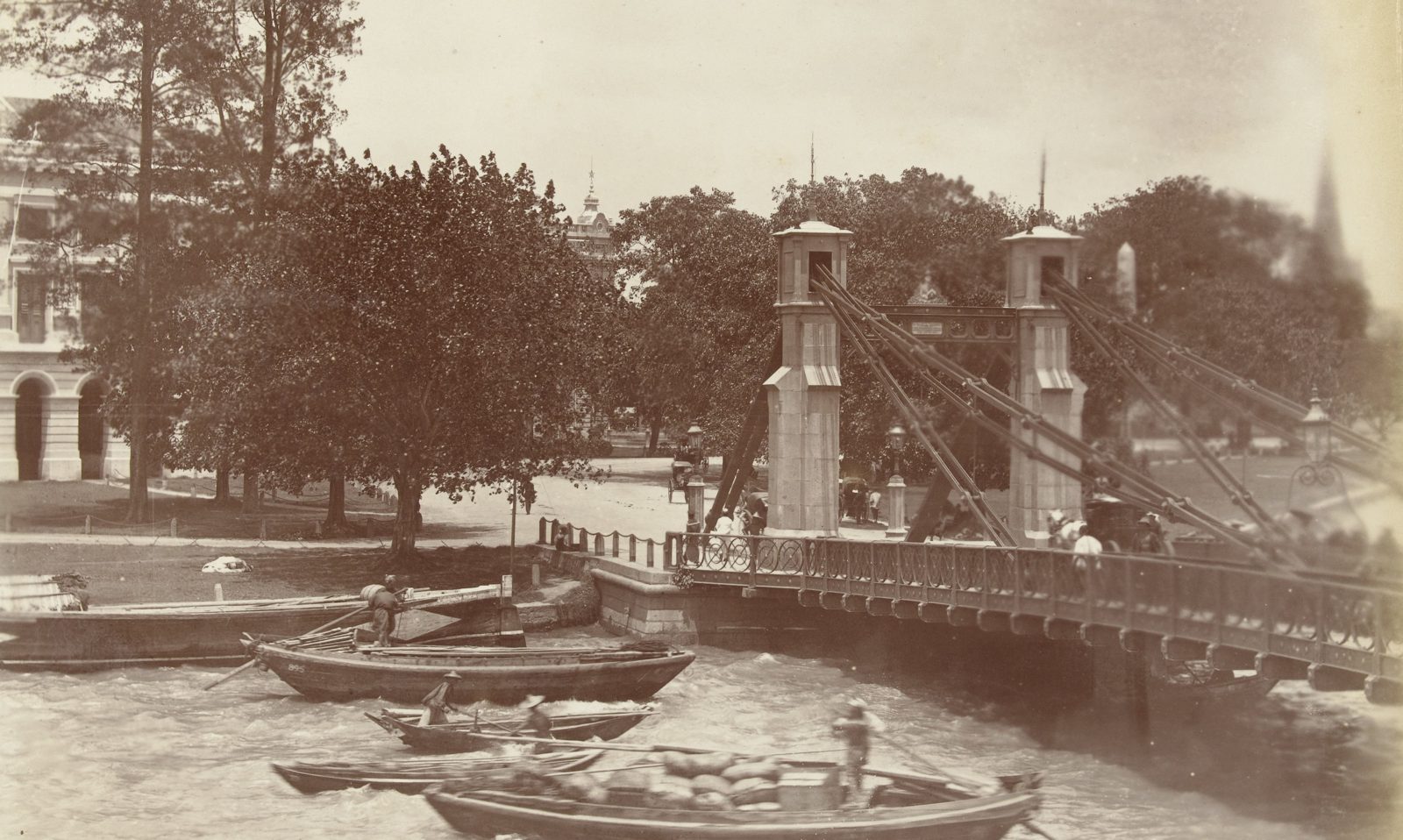 Wooden cargo boats loaded with sacks on the Singapore River beside the Cavenagh Bridge, with carriages crossing the bridge.