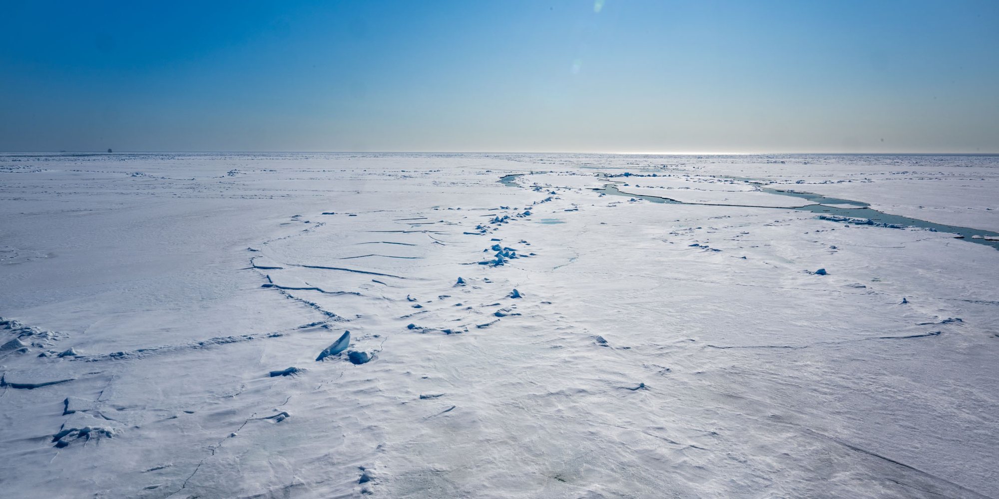Flat white sea ice stretching to the horizon under a clear blue sky, with a lead of open water winding through it.