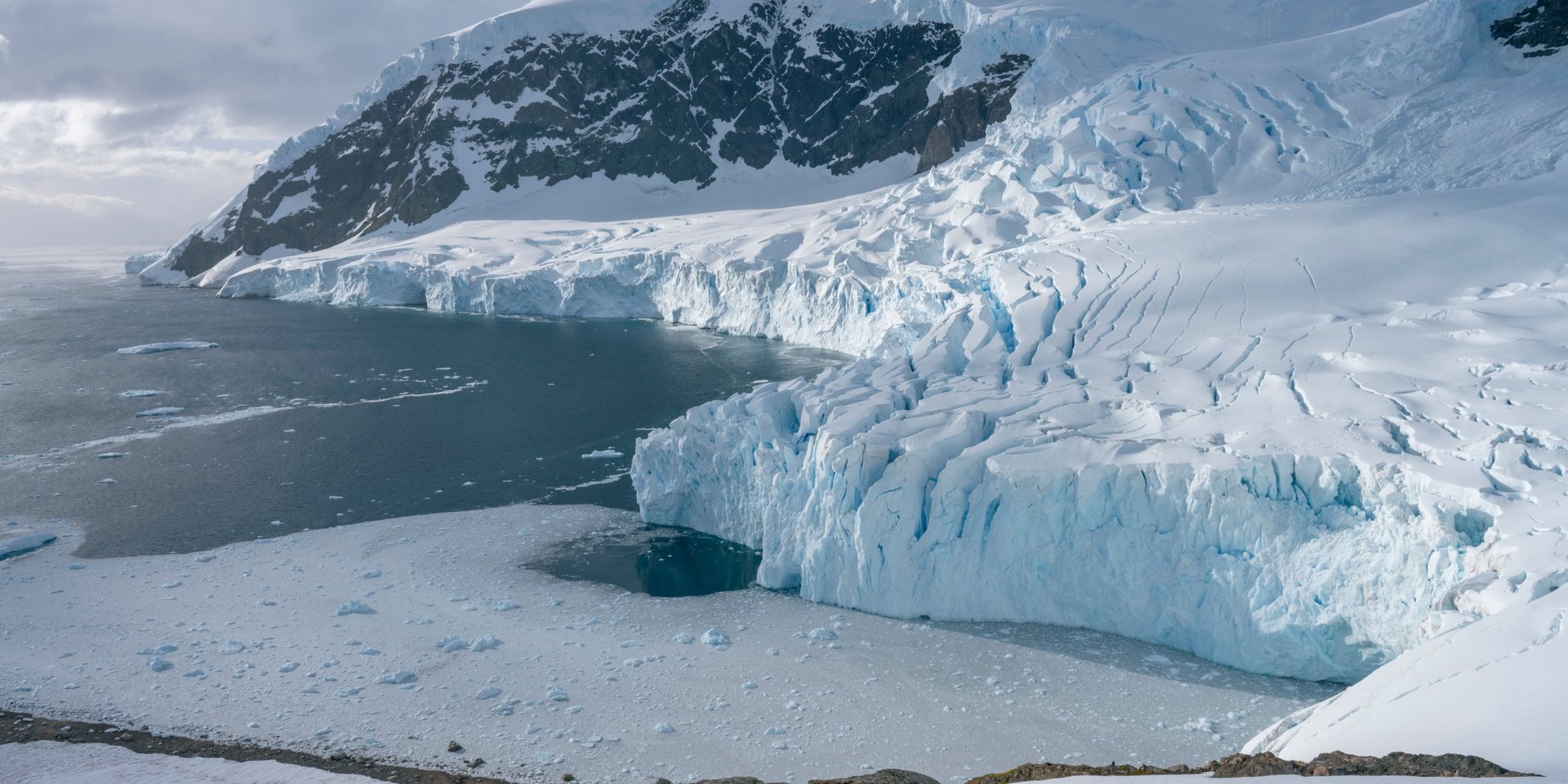 A glacier flowing down from a snow-covered mountain and ending in a cracked ice cliff at the edge of a dark bay dotted with brash ice.