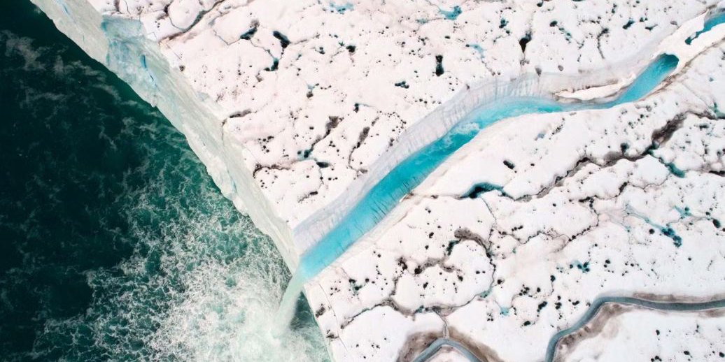 Seen from above, the edge of an ice cap: a turquoise meltwater river runs across the snow and pours off the ice wall into a dark green sea.