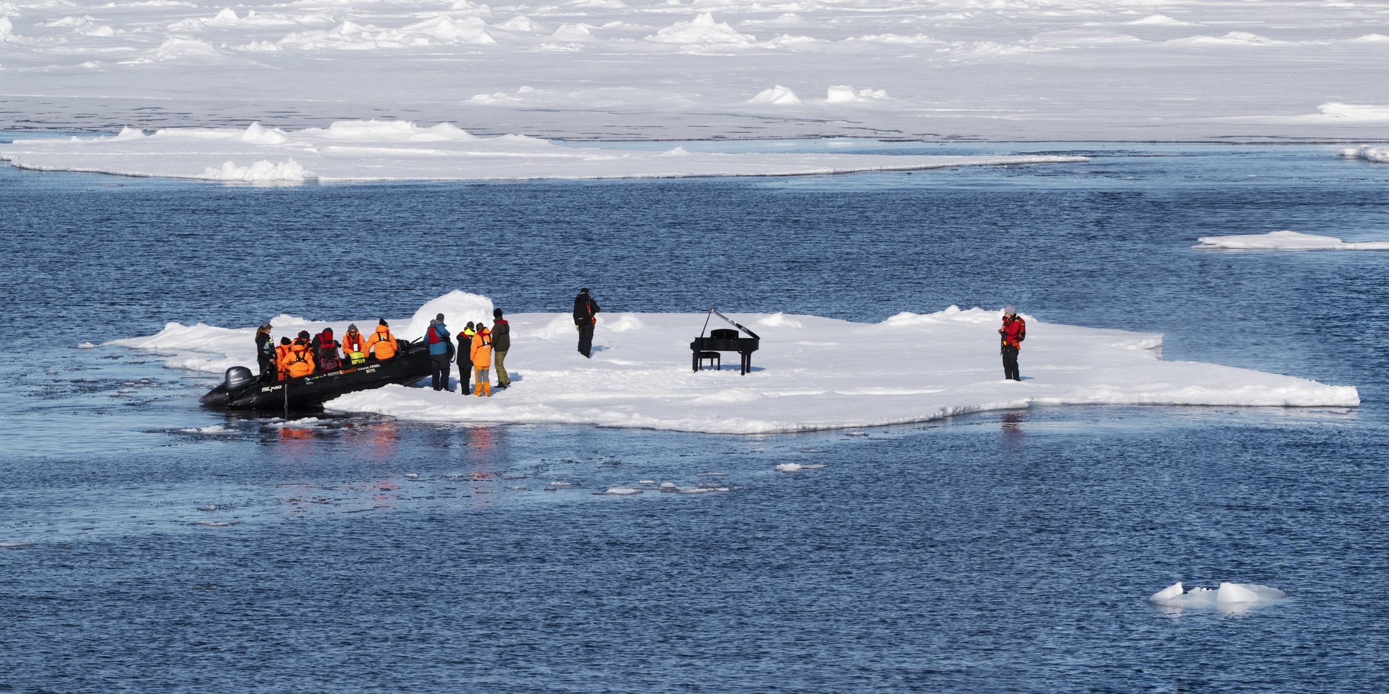 A black grand piano standing on a floe of sea ice, with a Zodiac of people in orange life vests at one end and a figure in red at the other, open water all around.