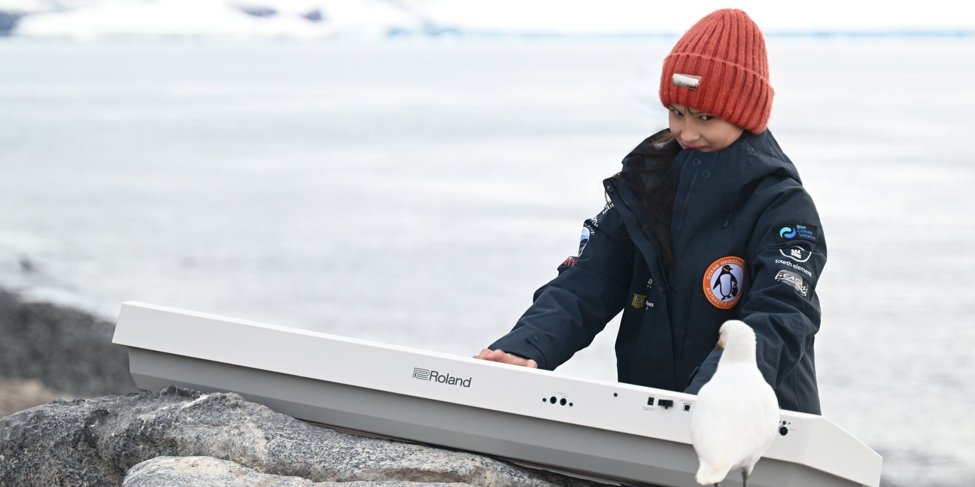 Hillary O’Sullivan, in an orange beanie and expedition jacket, playing a white keyboard on the rocks, with a white seabird standing on the end of the keyboard.