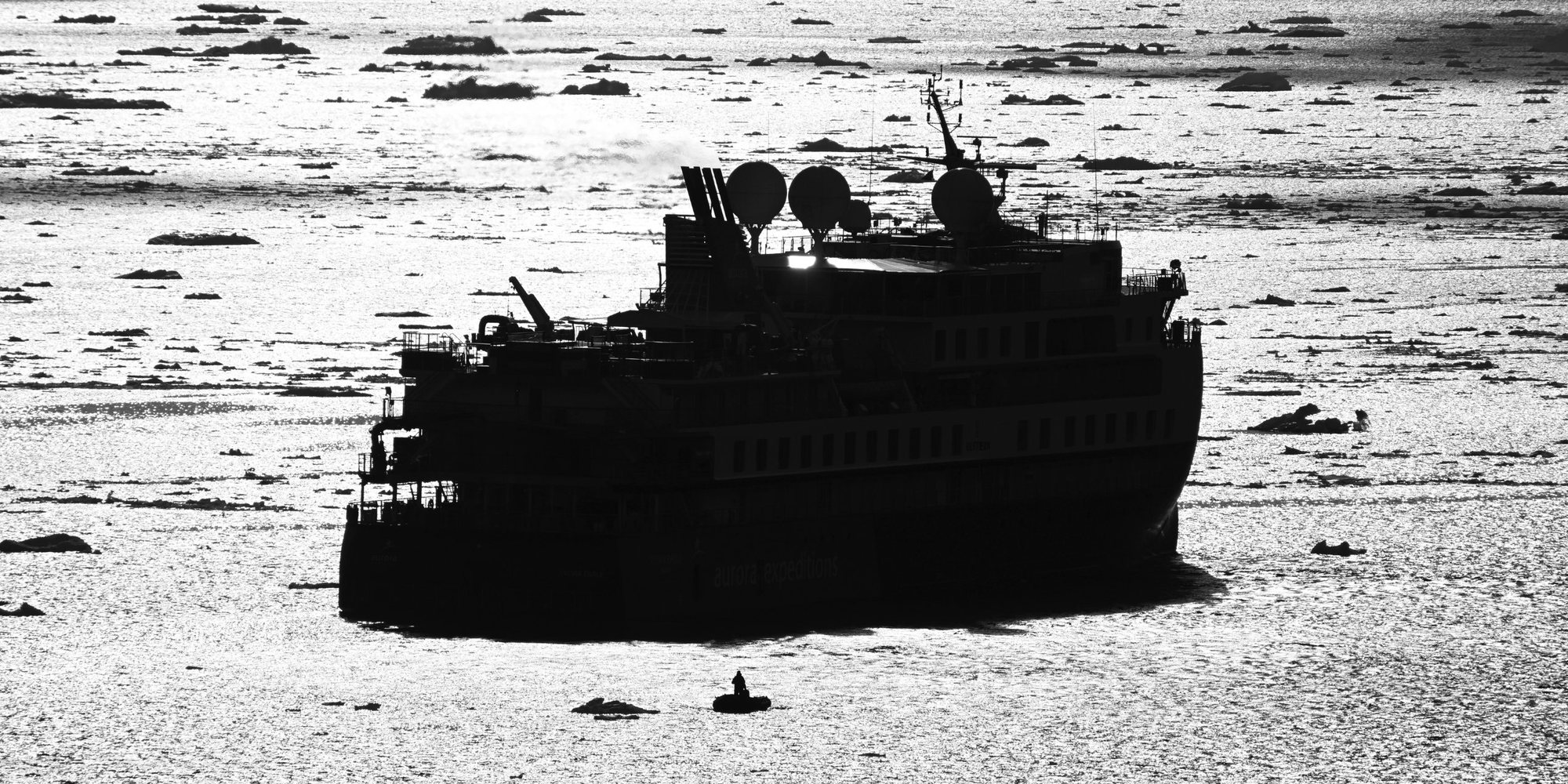 An expedition ship in silhouette against sunlit water scattered with ice floes, with a single Zodiac and its driver in the foreground.